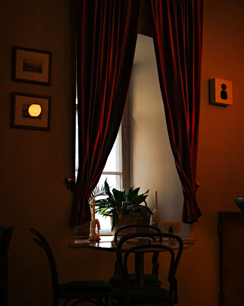 Fenêtre d’un café encadrée de rideaux rouges, chaises bistrot dans la pénombre.