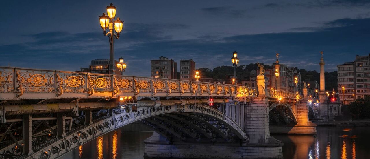Le pont de Fragnée à Liège à la tombée du jour, lampadaires dorés allumés au-dessus de la Meuse.